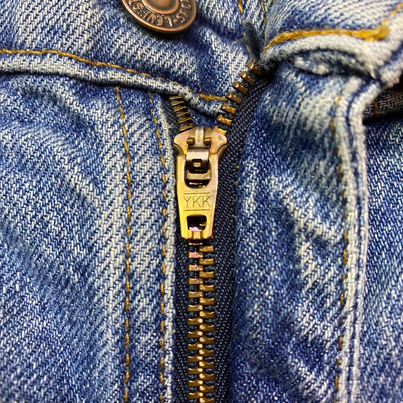 May include: Close-up of a blue denim garment with a brass-colored zipper. The zipper is partially closed, revealing the teeth and the YKK brand marking on the pull tab. The denim has visible stitching.