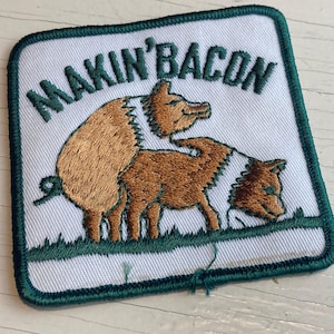 Peut inclure: Écusson brodé avec l'inscription "MAKIN' BACON" au-dessus d'un dessin de trois cochons bruns. L'écusson a un fond blanc avec une bordure verte et du texte vert. Les cochons sont représentés sur un patch avec de l'herbe verte.