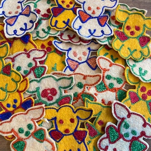 Peut inclure: Une collection d'appliqués en forme de chien en feutre de différentes couleurs. Les appliqués présentent des chiens jaunes, blancs et crème avec des accents rouges, verts et bleus. Chaque chien a un nœud papillon et est entouré d'un contour bleu. Idéal pour le bricolage.