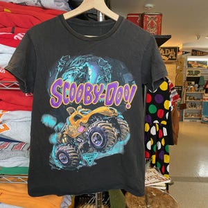 May include: Black t-shirt featuring a graphic of Scooby-Doo and a monster truck. The design includes the text "Scooby-Doo!" in purple and yellow, with a monster truck in yellow and blue. The shirt is hanging on a wooden hanger.