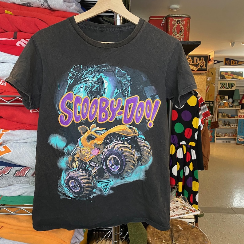 May include: Black t-shirt featuring a graphic of Scooby-Doo and a monster truck. The design includes the text "Scooby-Doo!" in purple and yellow, with a monster truck in yellow and blue. The shirt is hanging on a wooden hanger.