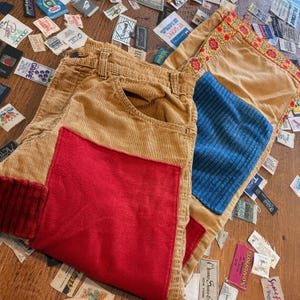 May include: Tan corduroy trousers with a red patch and turned-up cuff, next to a tan corduroy cushion with a blue patch and floral embroidery. Clothing labels are scattered around.