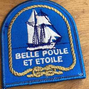 Peut inclure: Écusson brodé bleu avec un voilier blanc et des détails dorés. Le texte "BELLE POULE ET ETOILE" est brodé en blanc. De forme rectangulaire avec le haut arrondi, idéal pour personnaliser un vêtement ou un sac.