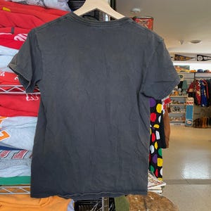 May include: A dark gray, short-sleeved t-shirt hanging on a wooden hanger. The shirt has a V-neck and appears to be made of a soft, worn material. The background shows a glimpse of a retail setting with various items.