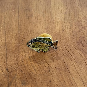 Könnte beinhalten: Ein detaillierter Fisch-Emaille-Pin mit gelben, grünen und blauen Farbtönen. Der Pin hat ein rotes Auge und einen silbernen Umriss. Der Pin befindet sich auf einem Holzgrund.