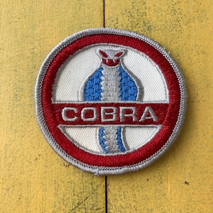 May include: Circular embroidered patch with a red ring and a grey border. The design features a blue and grey cobra on a white background, with the word "COBRA" in red. The patch has a stitched edge.
