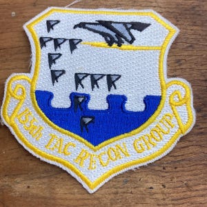 May include: Embroidered patch with a shield design. The patch features a white background with a blue lower section and a yellow border. The design includes a stylized bird, flags, and the text "85th TAC RECON GROUP".