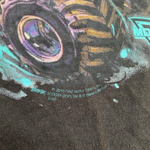 May include: Black t-shirt featuring a monster truck design with a large purple and gold wheel. The design includes teal accents and the text "MONSTER JAM". The shirt also has text that reads "&copy; 2018 Feld Motor Sports, Inc."