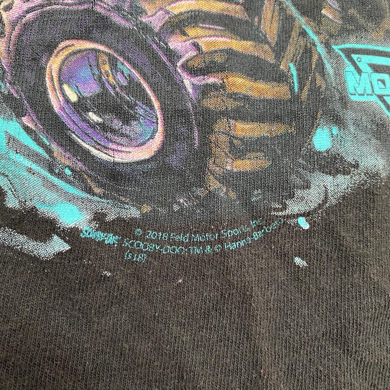 May include: Black t-shirt featuring a monster truck design with a large purple and gold wheel. The design includes teal accents and the text "MONSTER JAM". The shirt also has text that reads "&copy; 2018 Feld Motor Sports, Inc."