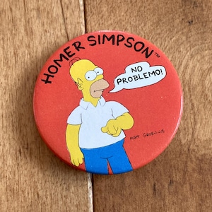 Peut inclure: Un bouton rond jaune et orange représentant Homer Simpson avec le texte "Homer Simpson" et "No Problemo!" sur fond rouge.