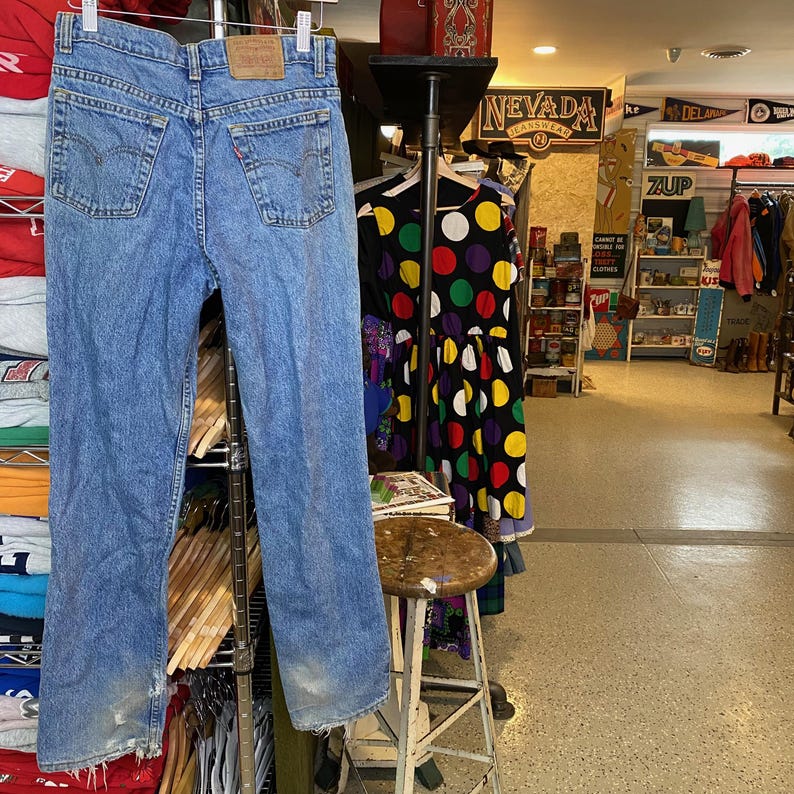 May include: Light blue denim jeans with a classic five-pocket design, hanging on a metal rack. The jeans show signs of wear and tear, with faded areas and distressed details. A brown leather patch is visible on the back. The jeans are a vintage style.
