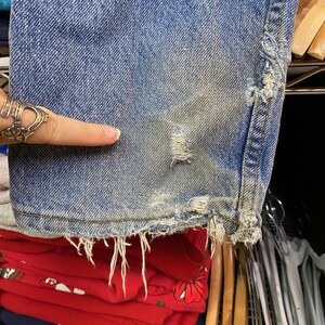 May include: Close-up of distressed blue denim jeans. The jeans have a worn appearance with frayed edges and a small tear. The denim is faded in areas, showing signs of wear and tear. The bottom hem is frayed, and the fabric has a vintage look.