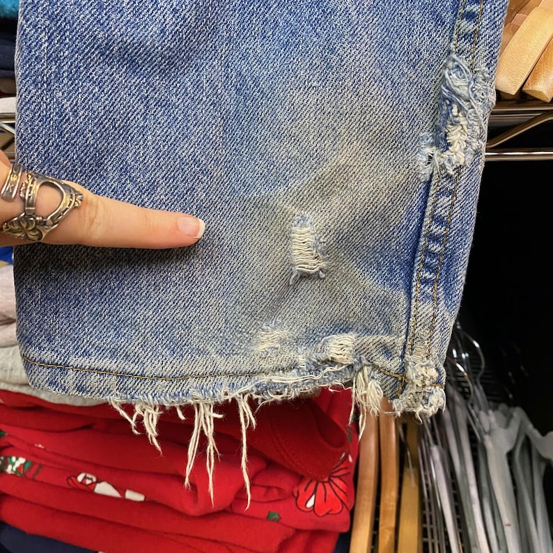 May include: Close-up of distressed blue denim jeans. The jeans have a worn appearance with frayed edges and a small tear. The denim is faded in areas, showing signs of wear and tear. The bottom hem is frayed, and the fabric has a vintage look.