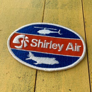 May include: Oval patch with a blue border and background, featuring a red horizontal stripe. The patch has white text that reads "Shirley Air", with a helicopter and airplane silhouette. The patch is likely for aviation enthusiasts.