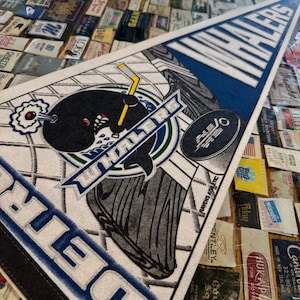 May include: A triangular pennant featuring the text "WHALERS" in white on a blue background. The pennant has a logo of a whale with a hockey stick and the text "WHALERS". The pennant is white with blue and black accents.