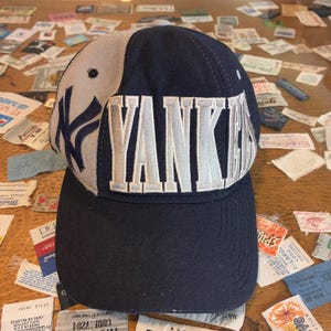 May include: A navy blue and grey baseball cap featuring the word "Yankees" embroidered in white. The New York Yankees logo is on the side. The cap has a curved peak and a structured crown.