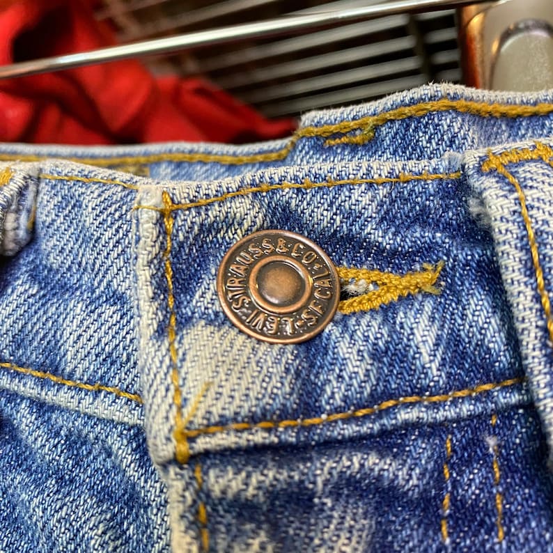May include: Close-up of a pair of blue denim jeans. The jeans feature a copper-colored button with the words "LEVI STRAUSS & CO. SF CAL." The jeans have yellow stitching and a faded appearance.