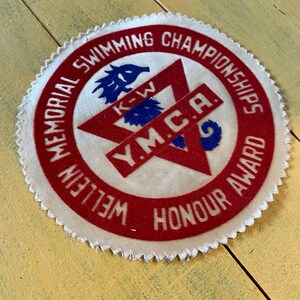 以下が含まれることがあります： 赤い縁とテキストが入った丸い白い布パッチ。「Memorial Swimming Championships Wellien Honour Award」と「Y.M.C.A.」の文字。中央には赤い星と青いエンブレムがあります。