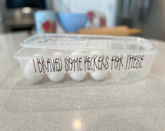 May include: A clear plastic egg carton with a lid, containing several white eggs. The front of the container features black text that reads, "I BRAVED SOME PECKERS FOR THESE," written in a stylized, thin, handwritten-style font.