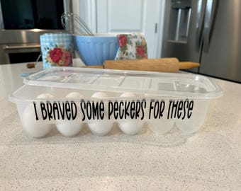 May include: A clear plastic egg storage container with a lid, holding six white eggs. The front of the container features black text that reads, "I BRAVED SOME PECKERS FOR THESE." The container is rectangular with molded individual egg slots.