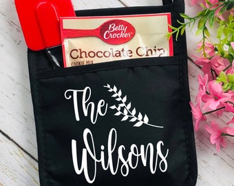 May include: Black oven mitt with a pocket containing a red spatula and a Betty Crocker Chocolate Chip cookie mix packet. The mitt features the text "The Wilsons" in white script with a decorative leaf design.