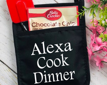 May include: Black oven mitt with the text "Alexa Cook Dinner" in white. A red spatula and a Betty Crocker Chocolate Chip cookie mix packet are in the pocket. The mitt has a loop for hanging.
