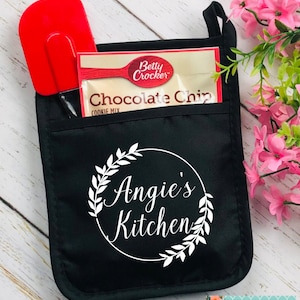 May include: Black oven mitt with a pocket containing a red spatula and a Betty Crocker Chocolate Chip cookie mix packet. The mitt features a white circular design with the text "Angie's Kitchen" and a floral wreath.