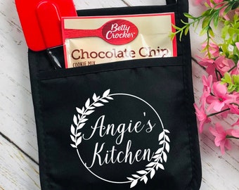 May include: Black oven mitt with a pocket containing a red spatula and a Betty Crocker Chocolate Chip cookie mix packet. The mitt features a white circular design with the text "Angie's Kitchen" and a floral wreath.