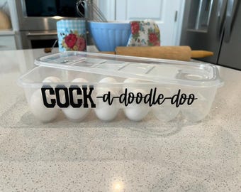 May include: A clear plastic egg storage container with a removable lid, holding five white eggs. The front of the container features black vinyl lettering that reads "COCK-a-doodle-doo" in a combination of block and script fonts.