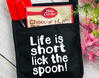 May include: Black oven mitt with the text "Life is short lick the spoon!" in white. A red spatula and a Betty Crocker chocolate chip cookie mix packet are in the pocket. A loop for hanging is at the top.
