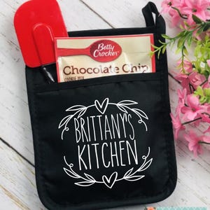 May include: Black oven mitt with the text "Brittany's Kitchen" in white script. A red spatula and a Betty Crocker Chocolate Chip Cookie Mix packet are in the pocket. A floral design is at the bottom right.