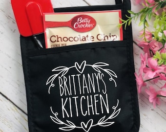 May include: Black oven mitt with the text "Brittany's Kitchen" in white script. A red spatula and a Betty Crocker Chocolate Chip Cookie Mix packet are in the pocket. A floral design is at the bottom right.