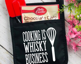 May include: Black pot holder with the text "COOKING IS WHISKY BUSINESS" and a white whisk graphic. A red spatula and a Betty Crocker chocolate chip cookie mix packet are in the pocket. The pot holder has a loop for hanging.