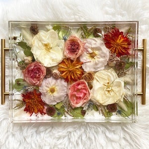 Op de afbeelding: Een transparante acryl dienblad met gouden handvatten gevuld met geperste bloemen. De bloemen zijn een mix van wit, roze, rood en oranje.
