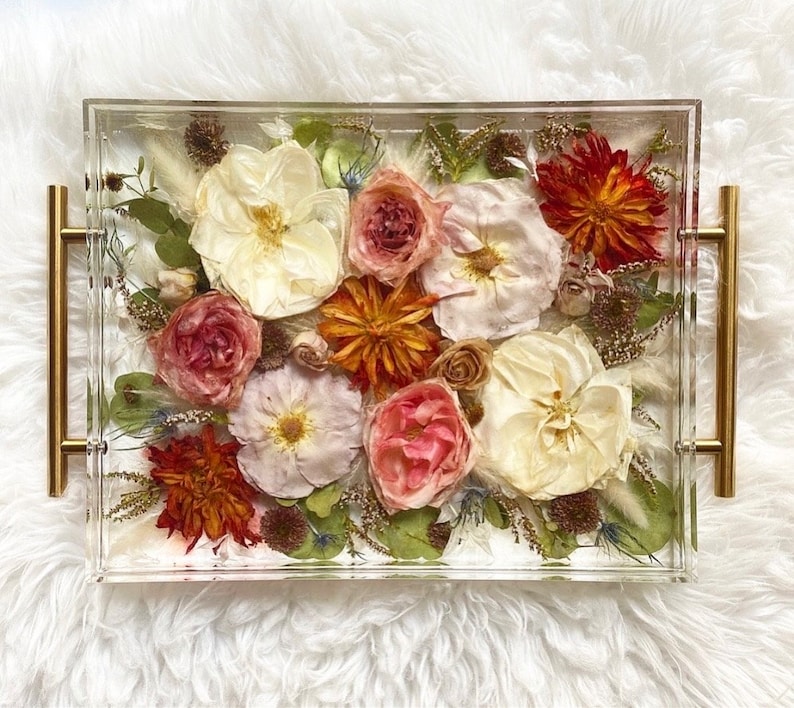 Op de afbeelding: Een transparante acryl dienblad met gouden handvatten gevuld met geperste bloemen. De bloemen zijn een mix van wit, roze, rood en oranje.