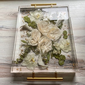 Op de afbeelding: Een transparante acryl dienblad met een gouden handvat, met een geperste bloemenarrangement van witte rozen, orchidee&euml;n en groen. De bloemen zijn geconserveerd in hars, waardoor een uniek en elegant display ontstaat.