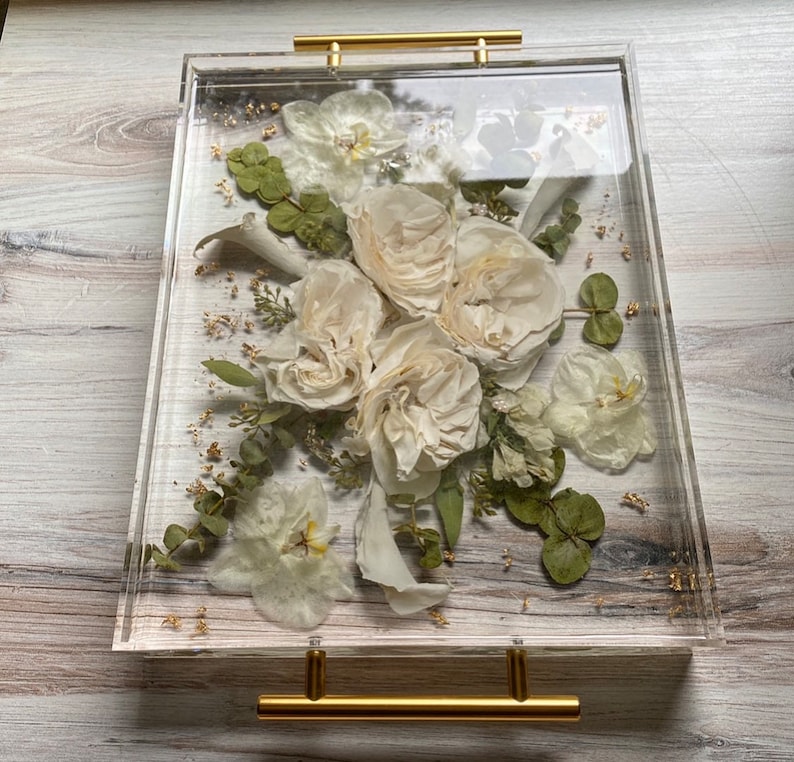 Op de afbeelding: Een transparante acryl dienblad met een gouden handvat, met een geperste bloemenarrangement van witte rozen, orchidee&euml;n en groen. De bloemen zijn geconserveerd in hars, waardoor een uniek en elegant display ontstaat.