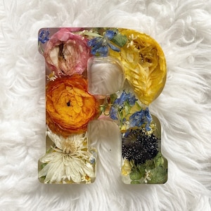 May include: A resin letter R with pressed flowers. The letter is white and the flowers are yellow, orange, pink, blue, and green.