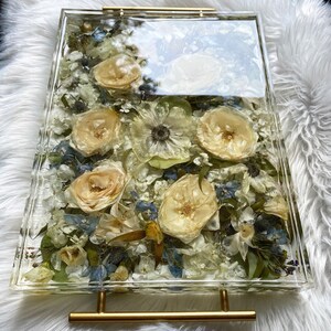 Op de afbeelding: Een transparante rechthoekige dienblad met een gouden metalen handvat. Het dienblad is gevuld met geperste bloemen en groen, waaronder witte rozen, blauwe bloemen en groene bladeren.