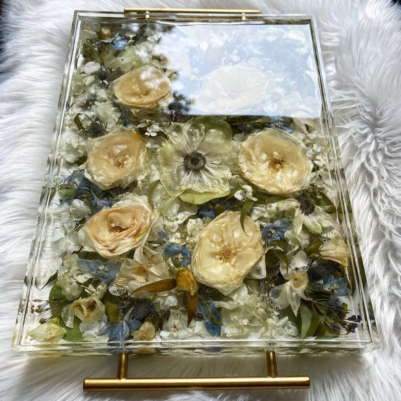 Op de afbeelding: Een transparante rechthoekige dienblad met een gouden metalen handvat. Het dienblad is gevuld met geperste bloemen en groen, waaronder witte rozen, blauwe bloemen en groene bladeren.