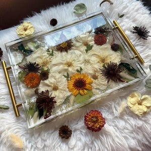 Op de afbeelding: Een transparante acryl dienblad met gouden handvatten gevuld met geperste bloemen en bladeren. De bloemen zijn wit, geel, oranje en bruin. Het dienblad ligt op een wit, pluche oppervlak.