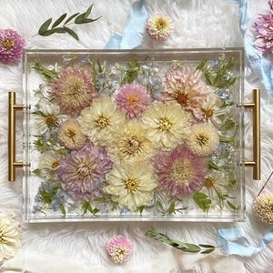 Op de afbeelding: Een rechthoekige, transparante dienblad met een gouden handvat aan elke kant. Het dienblad is gevuld met geperste bloemen in tinten roze, wit en geel. De bloemen zijn in de vorm van een boeket gerangschikt.