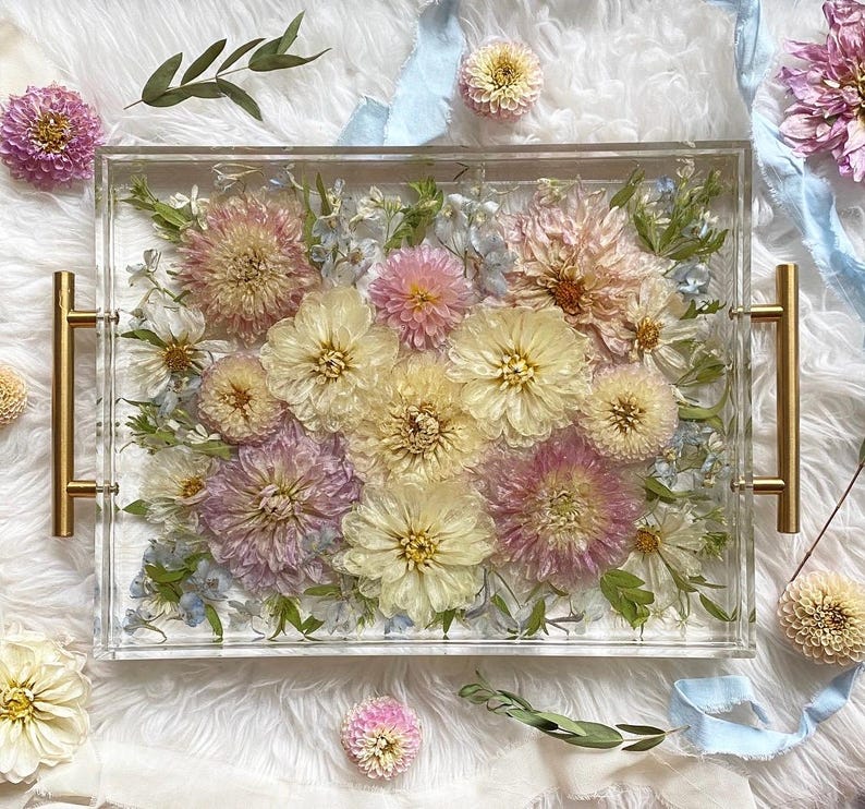 Op de afbeelding: Een rechthoekige, transparante dienblad met een gouden handvat aan elke kant. Het dienblad is gevuld met geperste bloemen in tinten roze, wit en geel. De bloemen zijn in de vorm van een boeket gerangschikt.