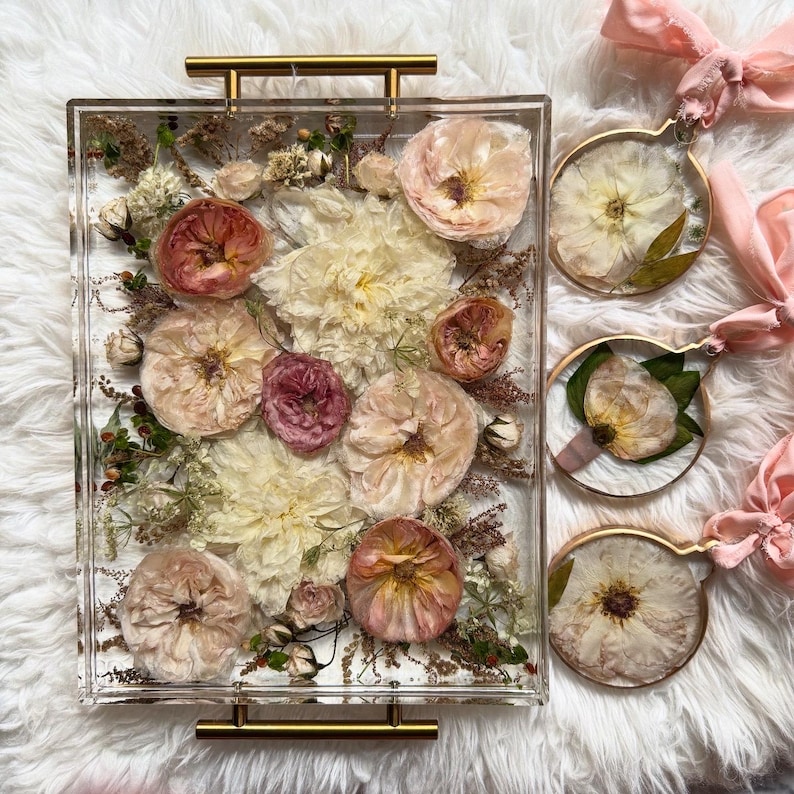 Op de afbeelding: Een transparante acryl dienblad gevuld met geperste bloemen en groen. Het dienblad heeft gouden metalen handvatten en is omgeven door drie kleinere ronde frames met geperste bloemen.