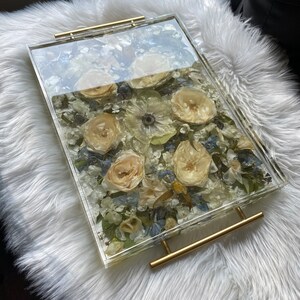 Op de afbeelding: Een transparante dienblad met een gouden handvat gevuld met geperste bloemen. De bloemen zijn voornamelijk wit en cr&egrave;me met enkele blauwe accenten.