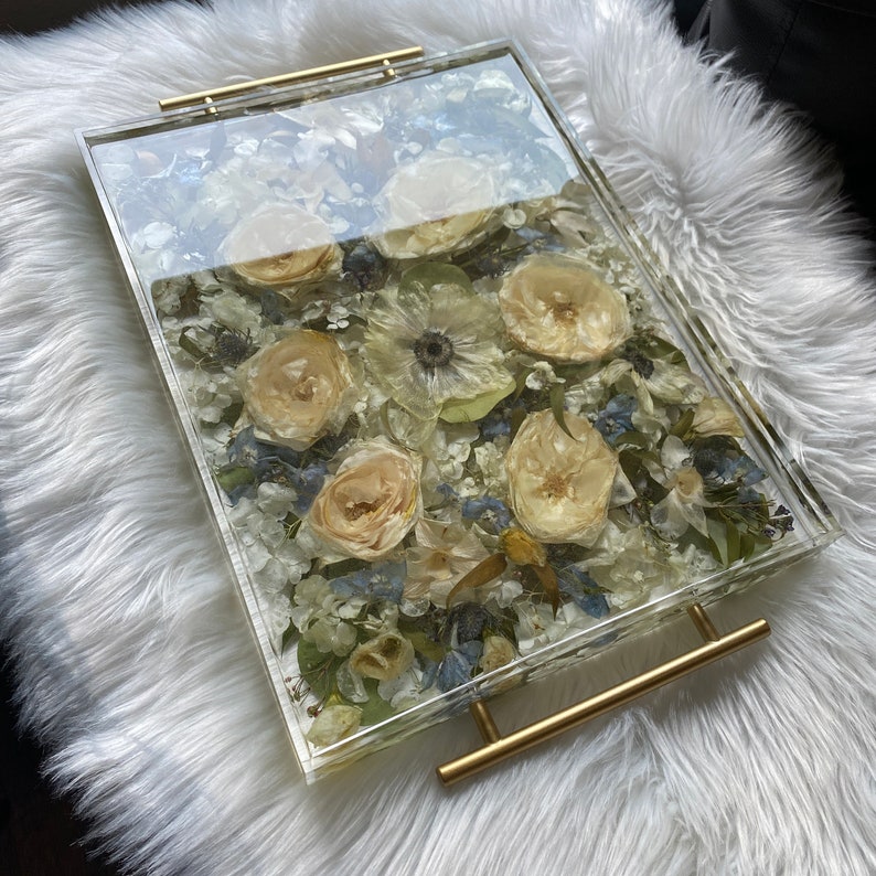 Op de afbeelding: Een transparante dienblad met een gouden handvat gevuld met geperste bloemen. De bloemen zijn voornamelijk wit en cr&egrave;me met enkele blauwe accenten.