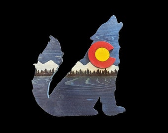May include: A wooden silhouette of a wolf howling at the moon, with a red, yellow, and white Colorado state flag design in the center. The wolf is painted blue and has a white mountain range and a row of trees behind it.
