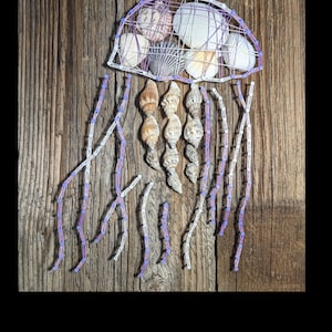 Könnte beinhalten: Eine lila und weiße String-Art-Qualle mit Muscheln. Die Qualle ist aus Nägeln gefertigt, die in ein Holzbrett geschlagen wurden, und mit Garn umwickelt. Die Muscheln dienen zur Gestaltung der Quallenhaube.