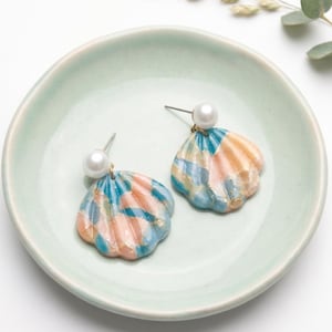 May include: A pair of stud earrings featuring shell-shaped pendants with a marbled pattern of blue, peach, and gold tones. Each earring is topped with a small, round white pearl bead attached to a metal post, creating a cohesive set of jewelry.