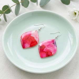 May include: A pair of leaf-shaped earrings with a vibrant, abstract design in shades of pink and red. The earrings have silver-colored hooks and are displayed on a light green plate. The earrings are a unique accessory.